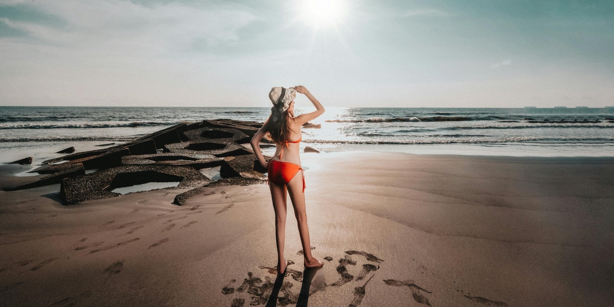 Žena u crvenom bikiniju stoji na plaži i gleda u more pod suncem, negovana koža iznutra zahvaljujući Premier Collagen.