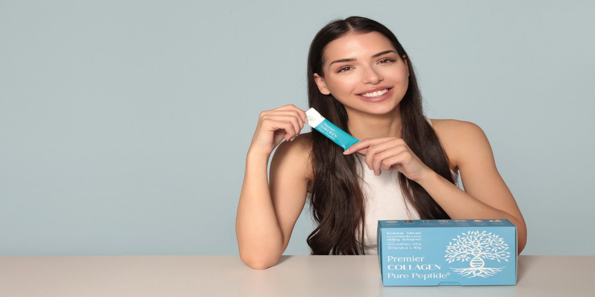 Nasmejana žena drži kesicu Premier Collagen Pure Peptide, sa kutijom proizvoda na stolu, prikazujući ambalažu i brending.